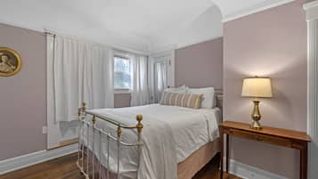 Un dormitorio con cama, una mesita de noche con lámpara, una ventana con cortinas y un cuadro en la pared.