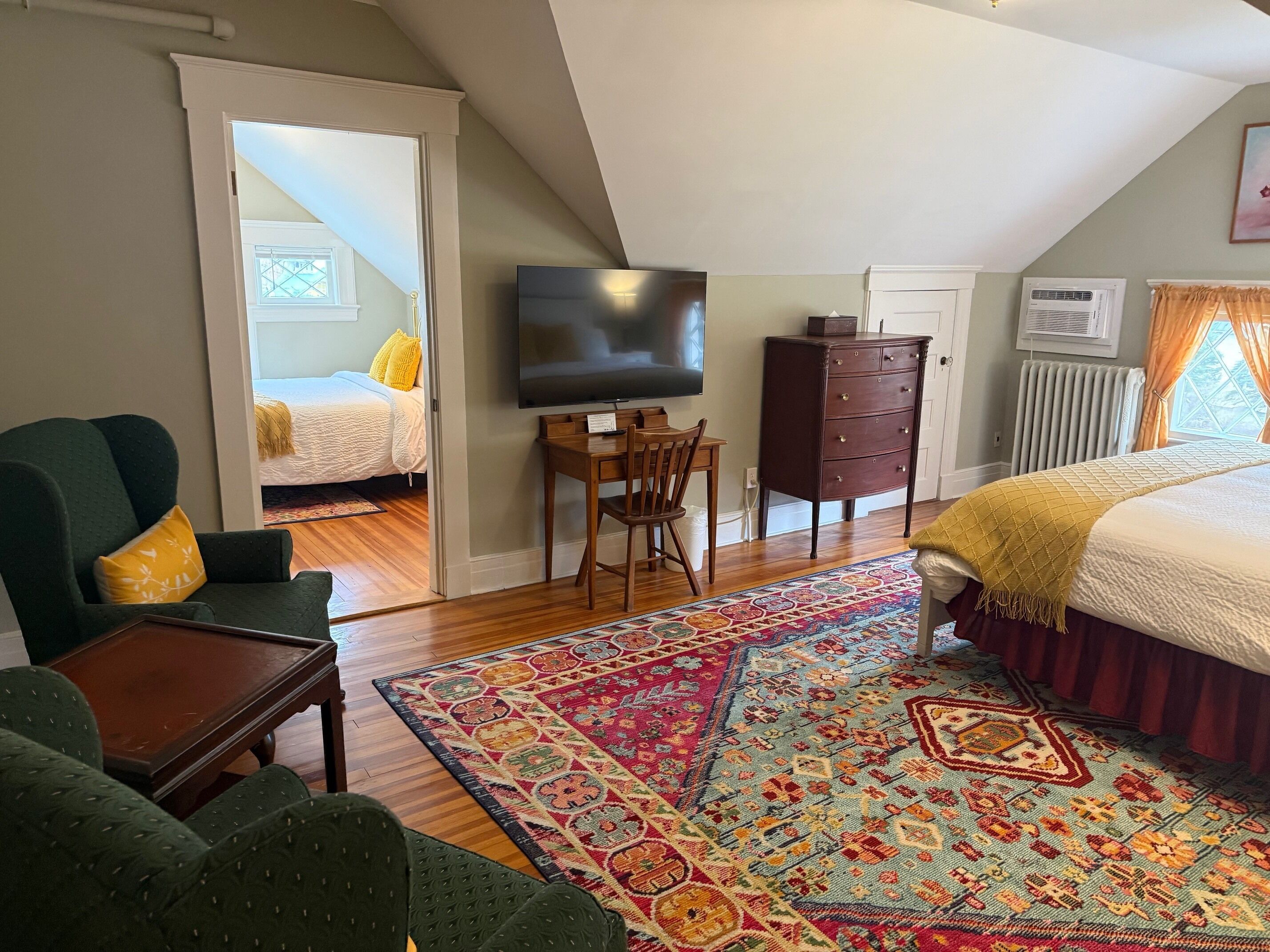 Un dormitorio con cama, televisión, escritorio, silla, cómoda y ventana con cortinas.