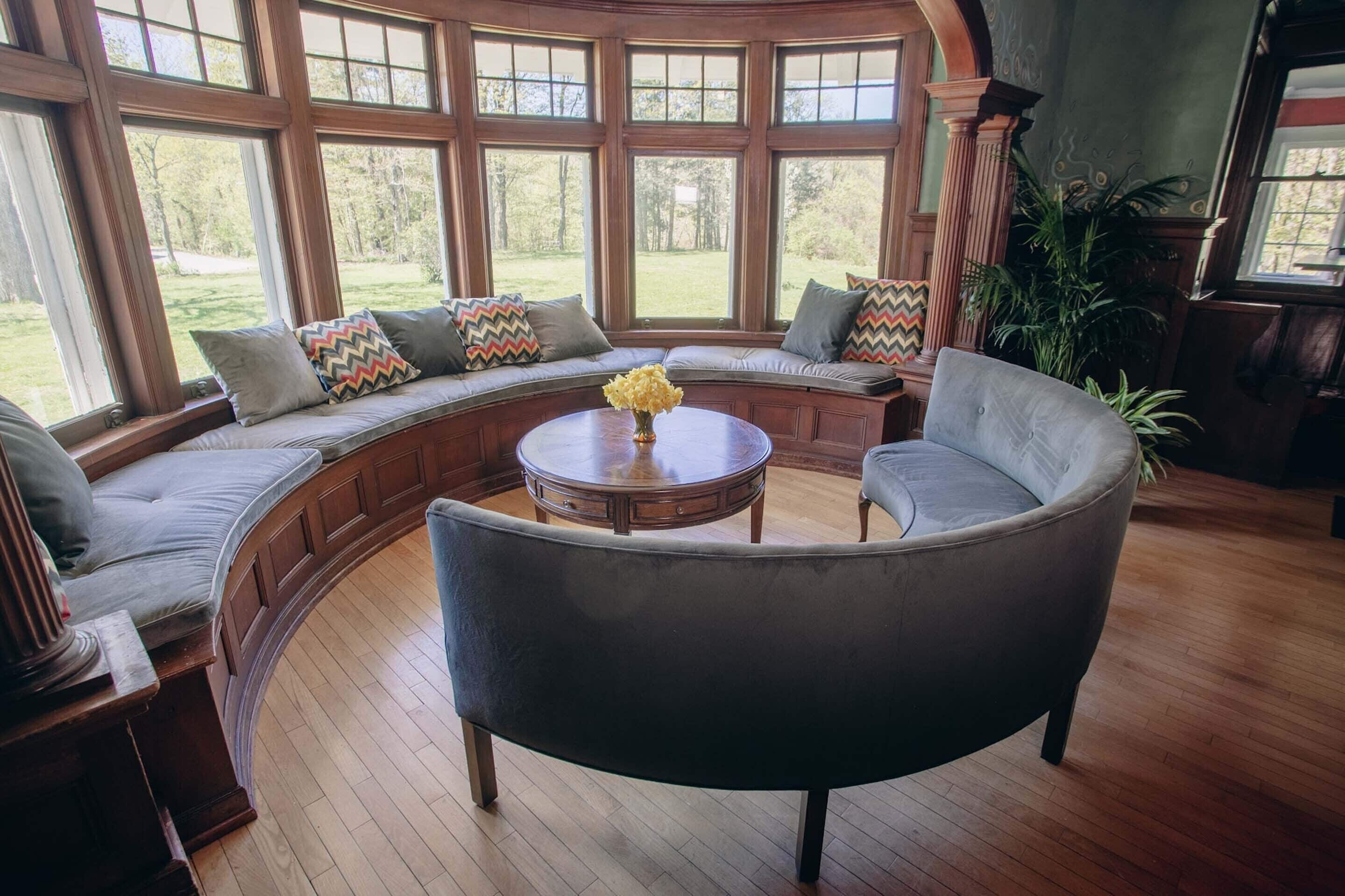 A cozy seating area with a curved bench and a round table, large windows offering a view of the outdoors, and a potted plant.