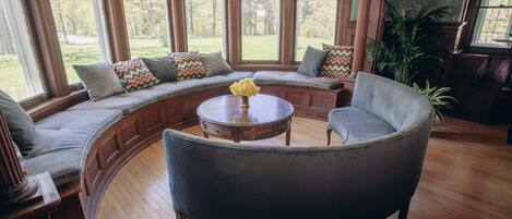A cozy seating area with a curved bench and a round table, large windows offering a view of the outdoors, and a potted plant.