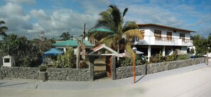 A two-story building with a stone wall, a blue roof, and a palm tree in front. - Hotel San Vicente Galapagos (Puerto Villamil)