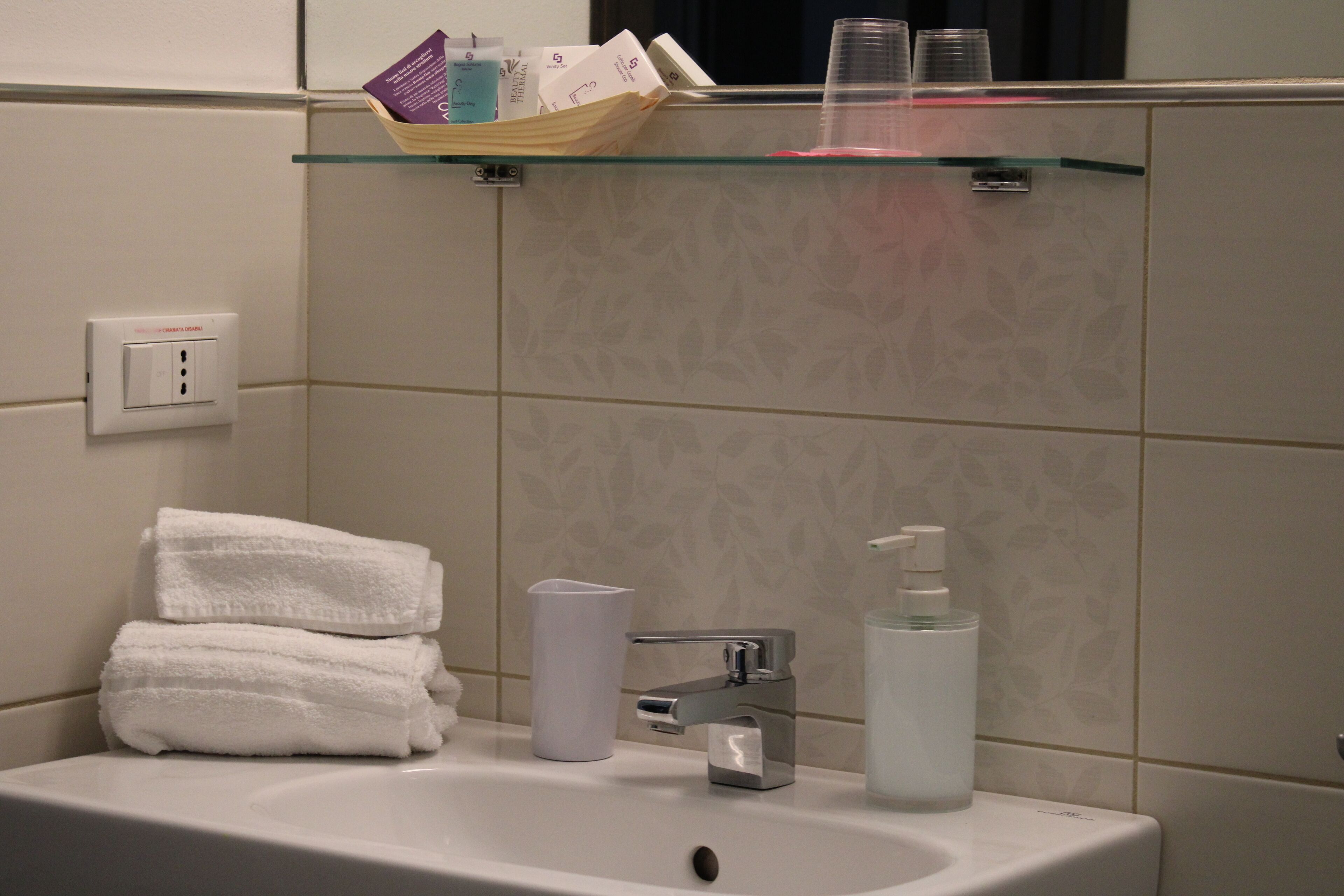 A bathroom with a sink, two stacked towels, a glass shelf with toiletries, and a mirror.