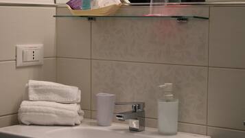 A bathroom with a sink, two stacked towels, a glass shelf with toiletries, and a mirror.