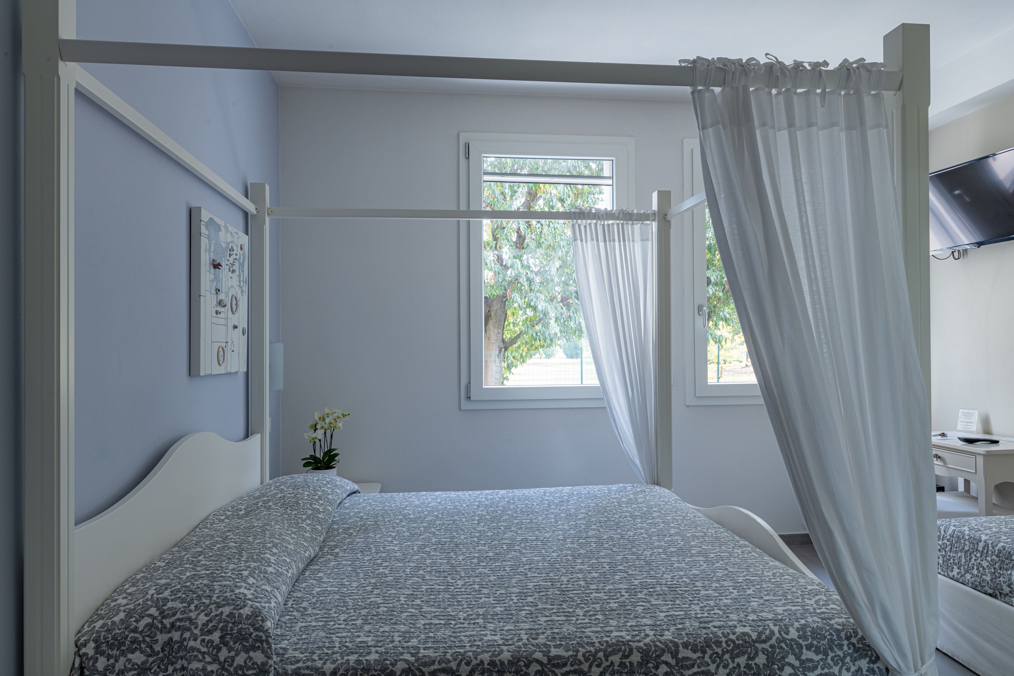 Un dormitorio con cama con dosel, un escritorio pequeño con silla, una ventana con vista a follaje y un televisor montado.