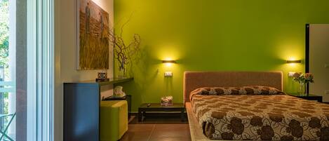 A modern bedroom with a green wall, a bed with a patterned bedspread, a bedside table, a nightstand, and a window with a view of greenery.