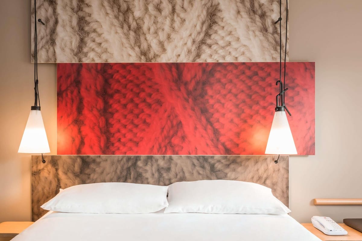A hotel room with a bed, two pendant lights, a red and beige textured wall art, a white pillow, and a bedside table with a phone.