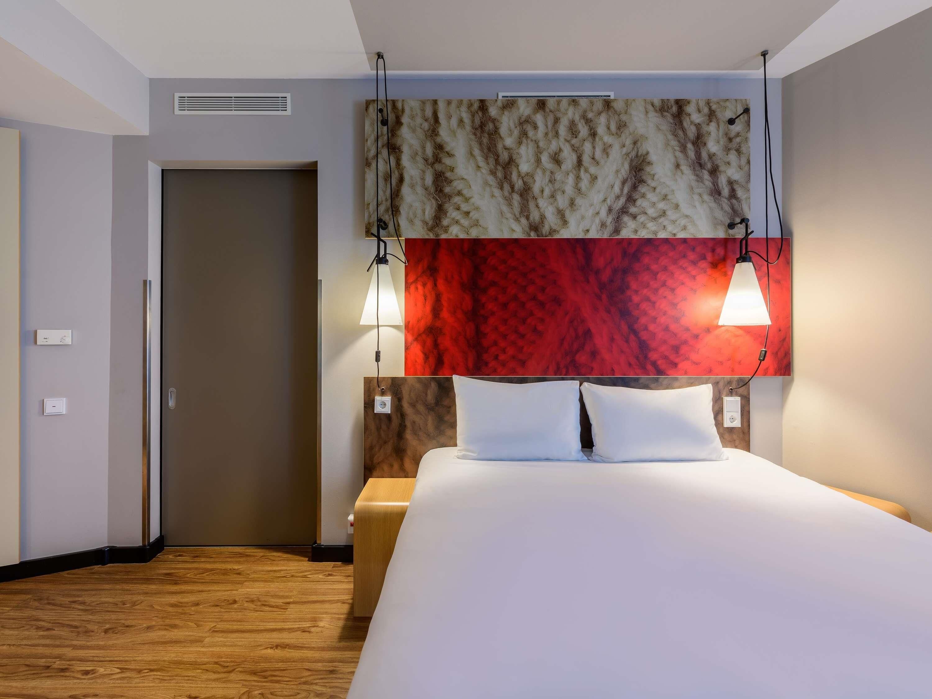 Habitación de hotel con cama, dos lámparas de noche, cabecera con diseño texturizado, suelo de madera y una puerta.