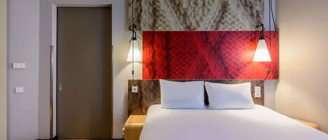 Habitación de hotel con cama, dos lámparas de noche, cabecera con diseño texturizado, suelo de madera y una puerta.