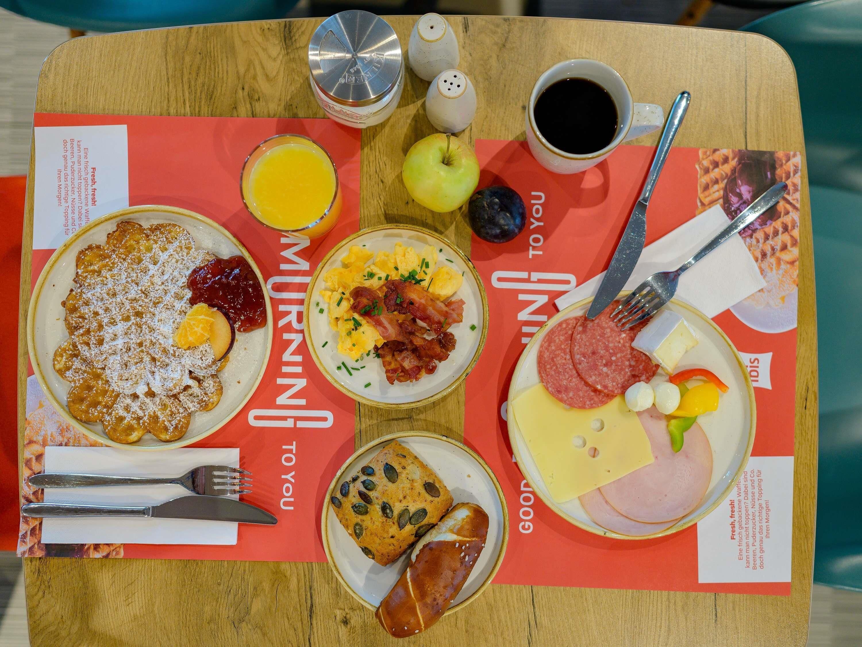 A breakfast tray with pancakes, scrambled eggs, bacon, ham, cheese, and a sausage on a wooden table.