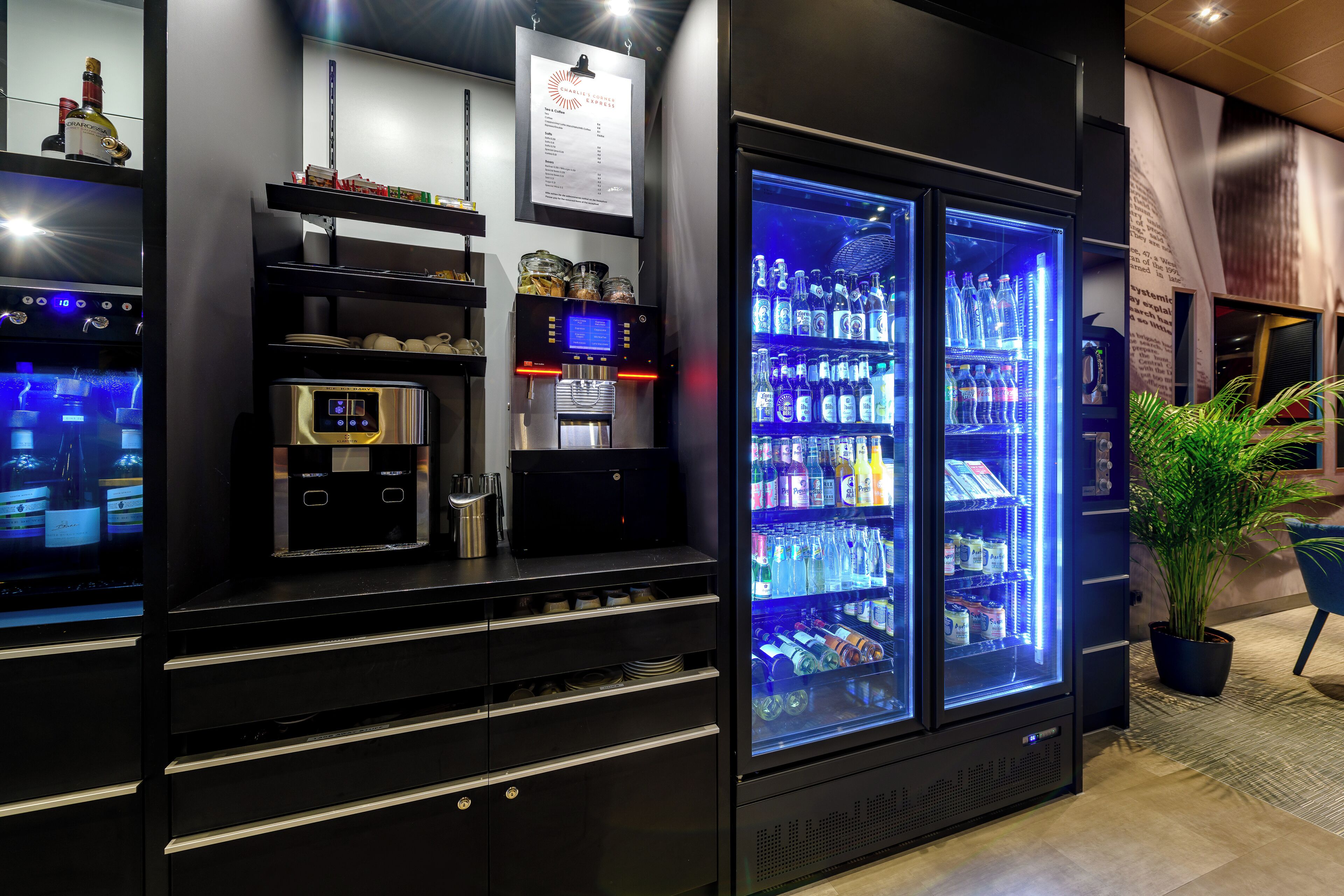 A minibar with a digital coffee maker, a refrigerator stocked with beverages, and a potted plant.