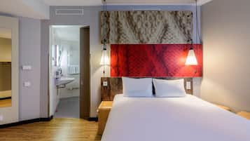 Una habitación de hotel moderna con una cama grande, suelo de madera, cabecero rojo y beige, y un baño visible a través de una puerta abierta.