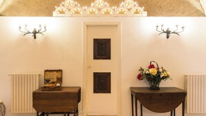 A room with a white door, a wooden table with a vase of flowers, a wooden chest, and wall sconces with lights. - Masseria Agrituristica Lama San Giorgio (Rutigliano)