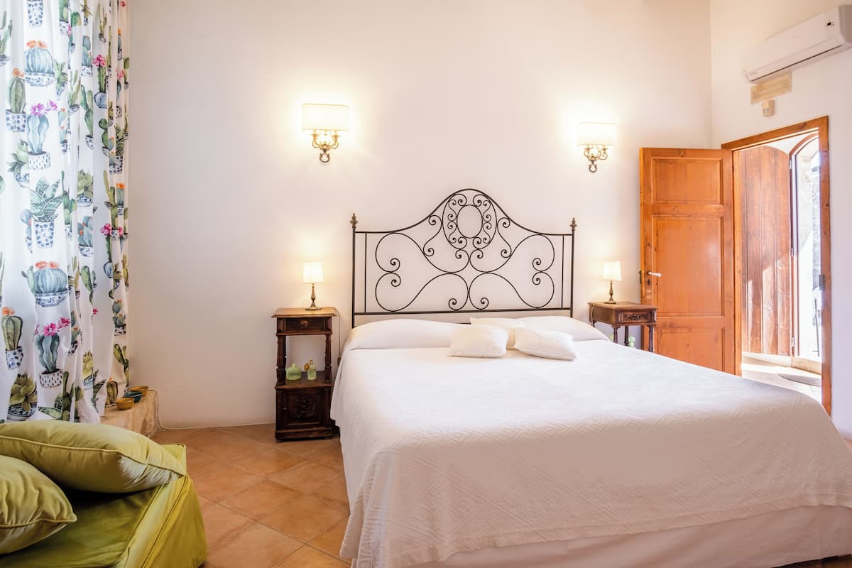 Un dormitorio con una cama, dos mesitas de noche, una mesilla auxiliar, una puerta de madera y una ventana.