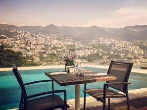 A poolside table with two chairs, overlooking a cityscape with buildings and hills. - Monte Cassino Boutique Hotel (Kfar Yassine)
