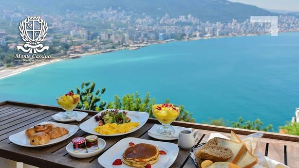 Una mesa con alimentos para el desayuno, como cruasanes, tortitas y pan, con vistas a un paisaje urbano costero.