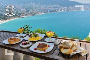 Taula amb menjar per esmorzar, com ara croissants, creps i pa, amb vista a una ciutat costanera.