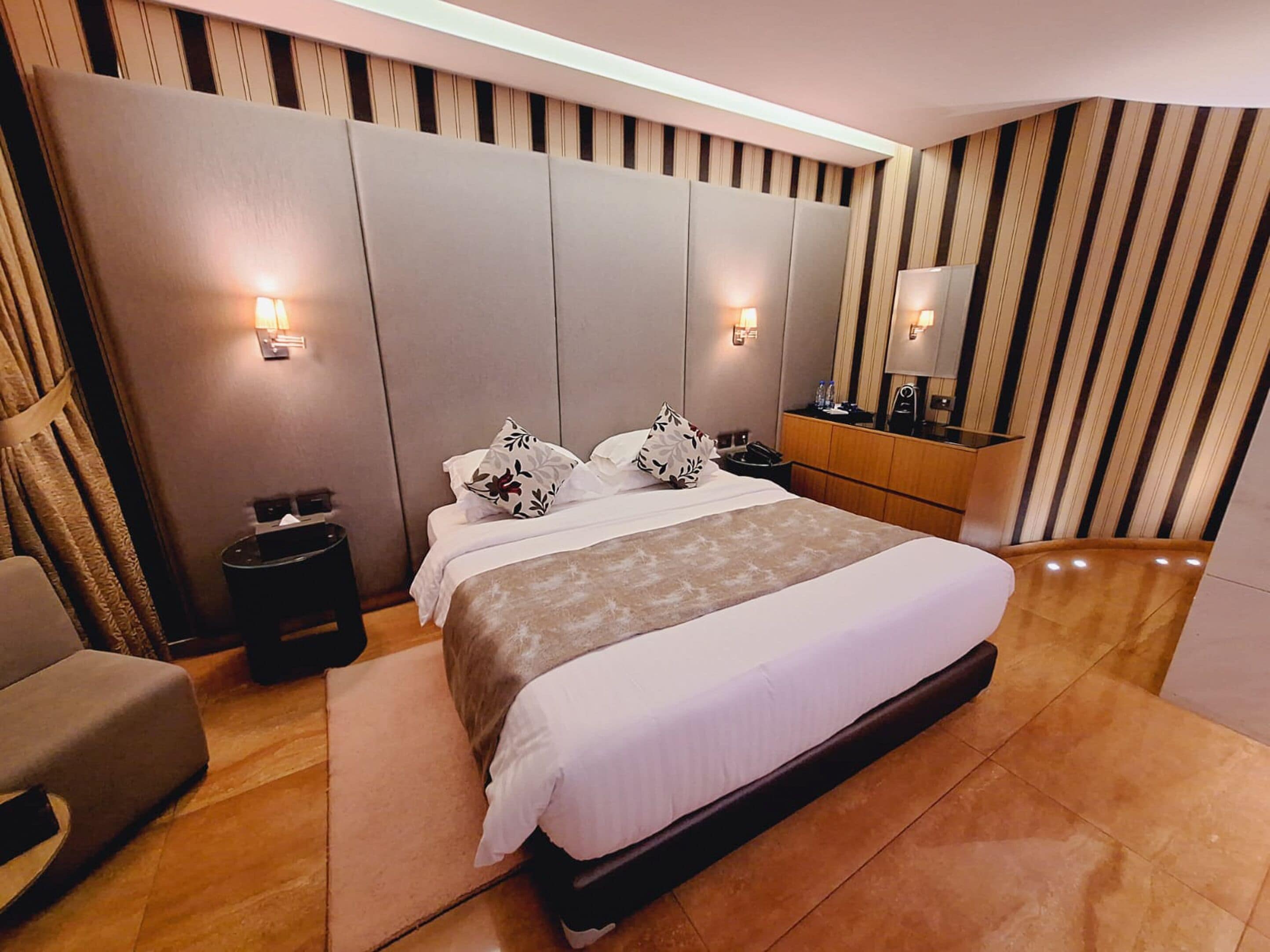 A modern hotel room with a large bed, a bedside table, a chair, a vanity with a mirror, and striped wallpaper.