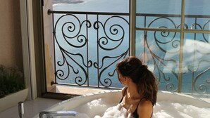 Una persona sentada en una bañera con vista a un paisaje urbano y una masa de agua a través de un ventanal grande.