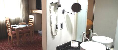 Habitación de hotel con zona de comedor, escritorio y silla, baño con lavamanos y espejo, y teléfono.