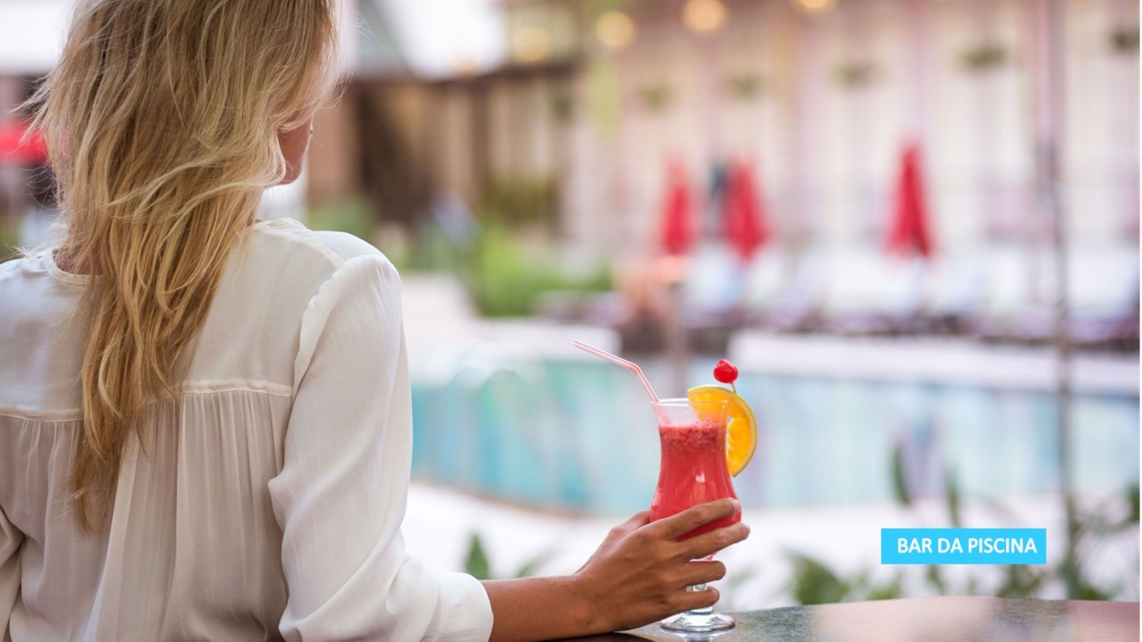 A person holding a cocktail with a straw and a cherry garnish, with a pool and umbrellas in the background.