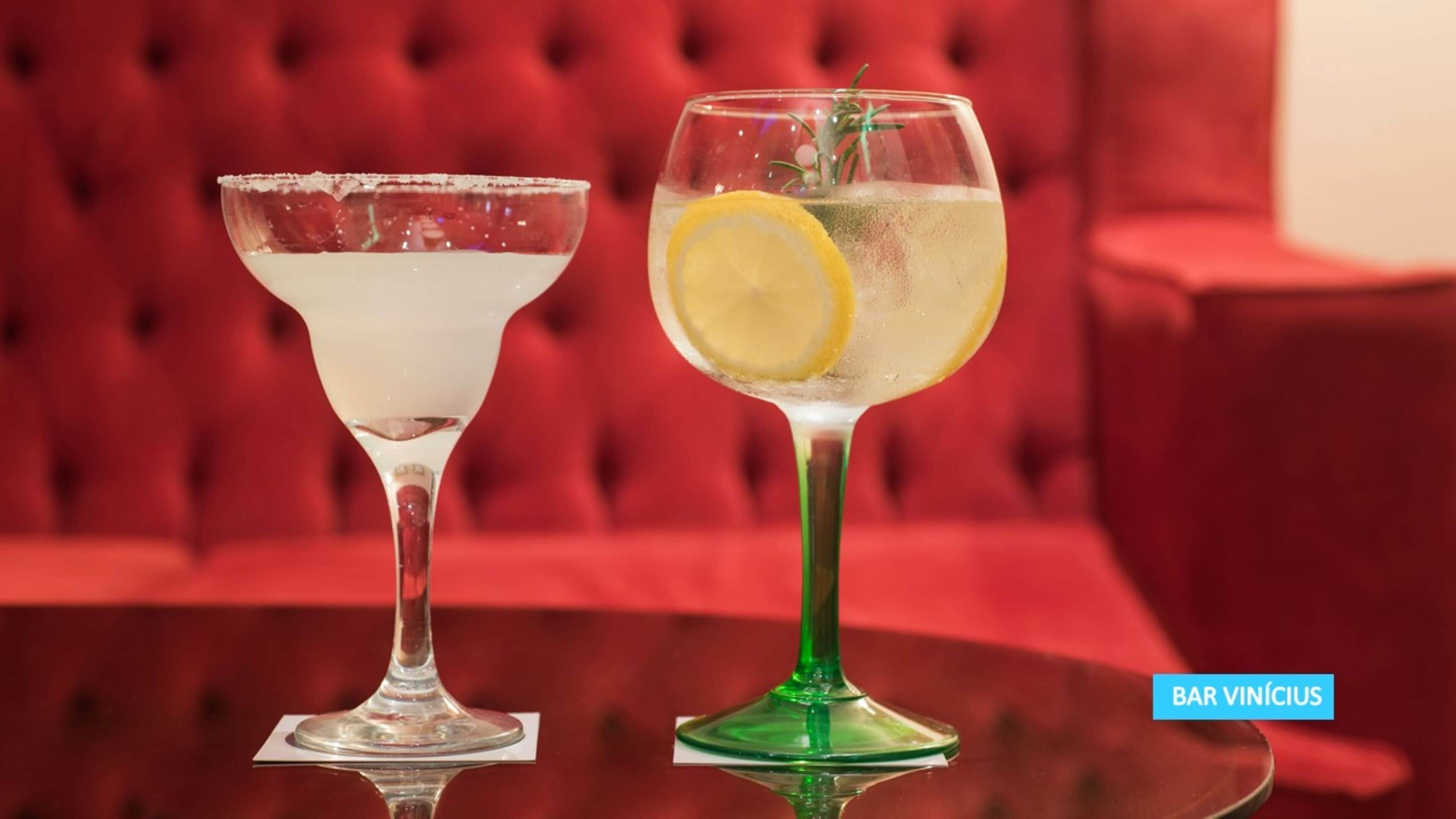 Due cocktail su un tavolo riflettente, con un divano imbottito rosso sullo sfondo.