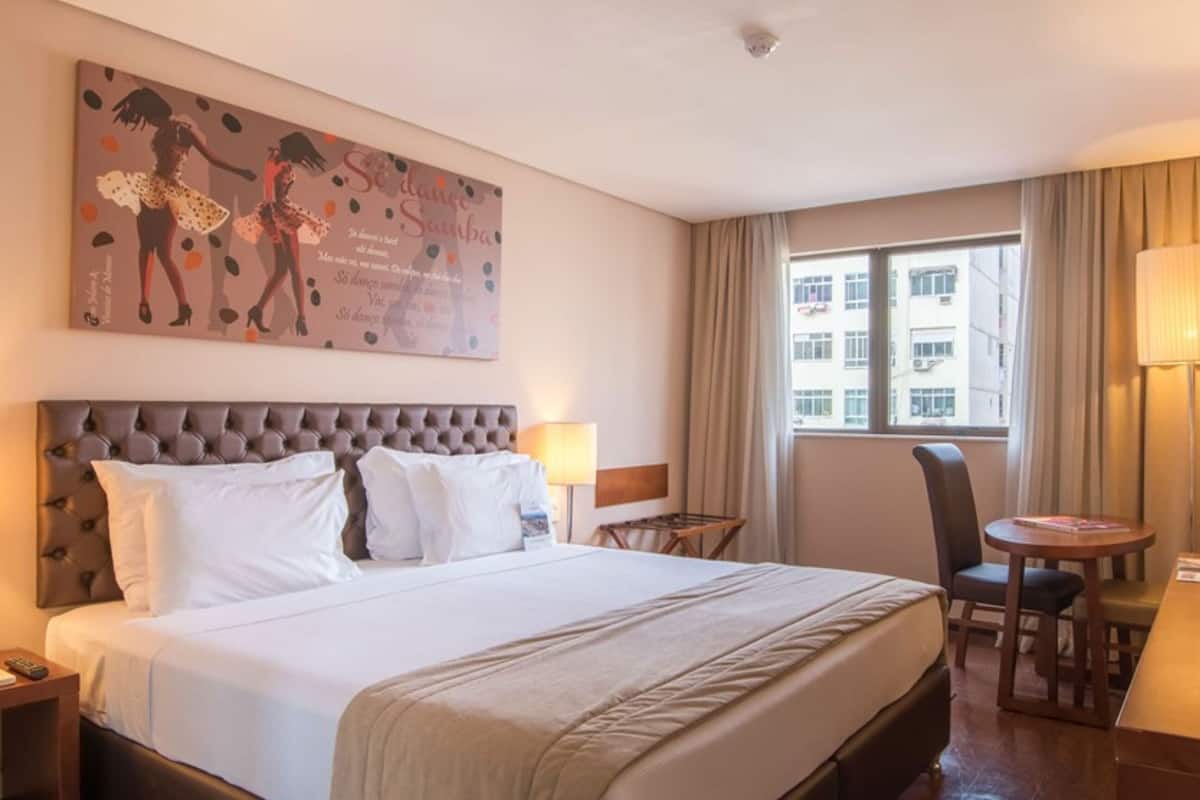 A hotel room with a large bed, two bedside lamps, a desk with a chair, a window with curtains, and a wall-mounted TV.