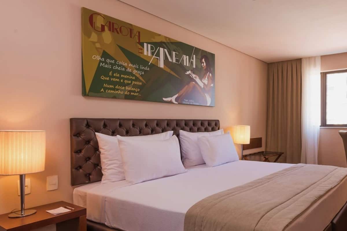 A hotel room with a large bed, two bedside lamps, a desk with a chair, a window with curtains, and a wall-mounted TV.