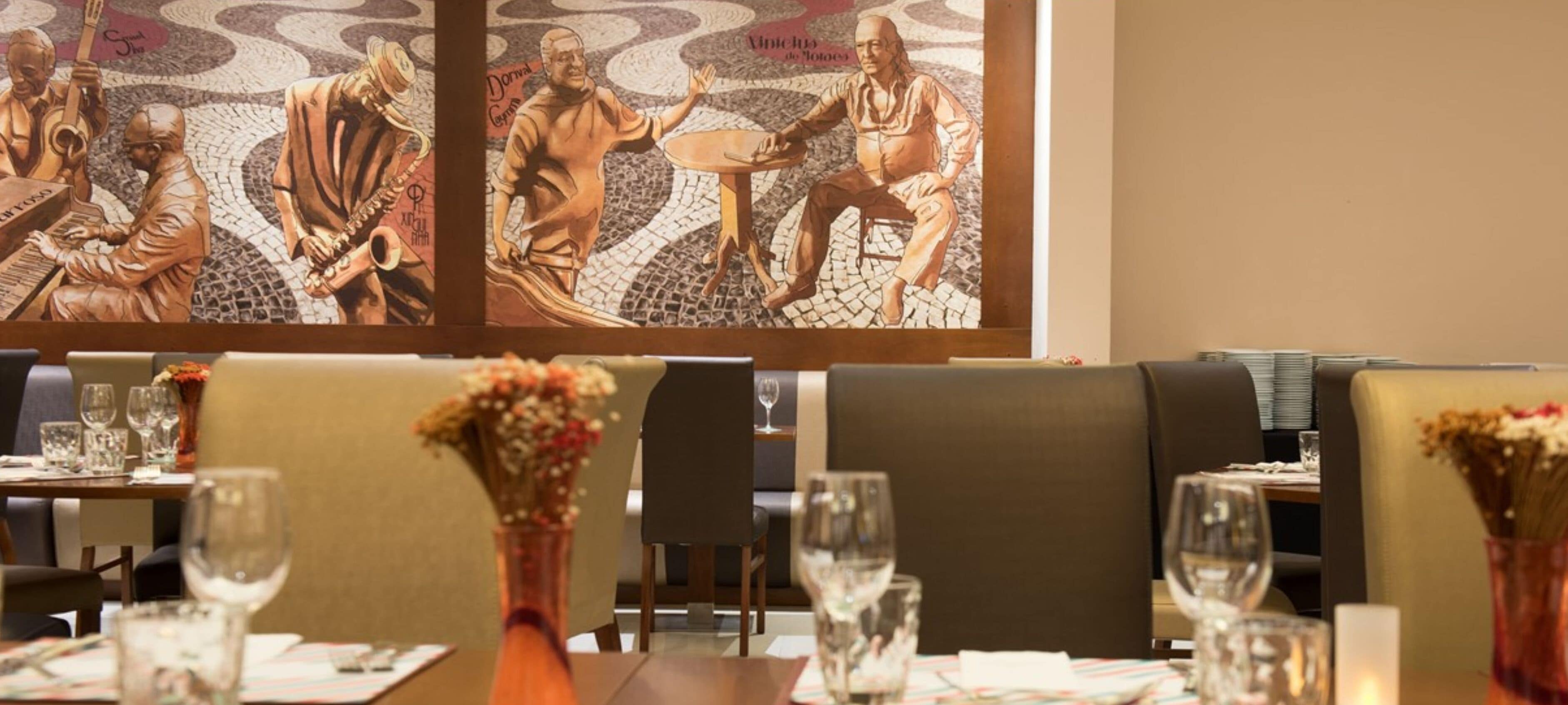A restaurant with tables set for dining, a mosaic wall art, and vases with flowers on the tables.