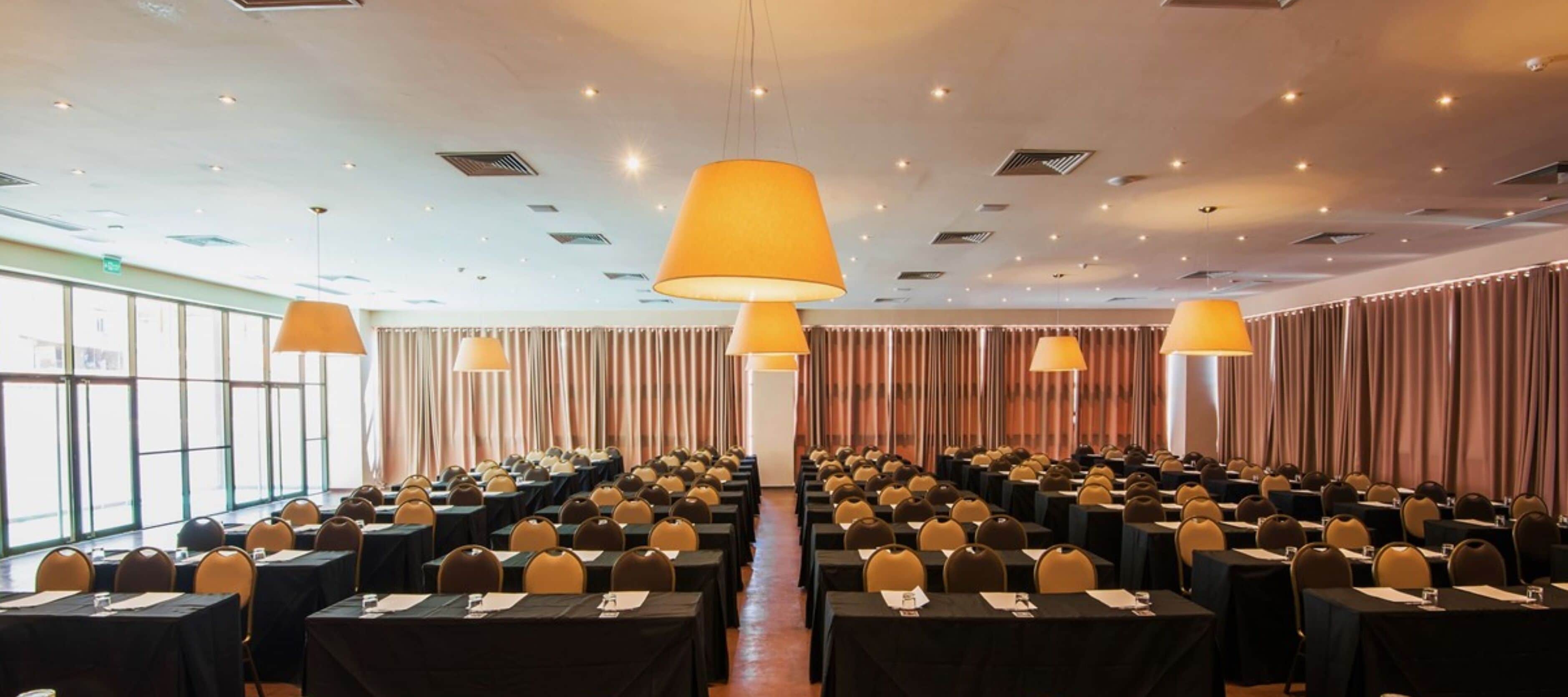 Un salón de conferencias con filas de mesas y sillas, lámparas grandes y un techo alto con iluminación empotrada.