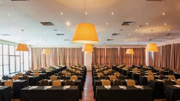 Un salón de conferencias con filas de mesas y sillas, lámparas grandes y un techo alto con iluminación empotrada.