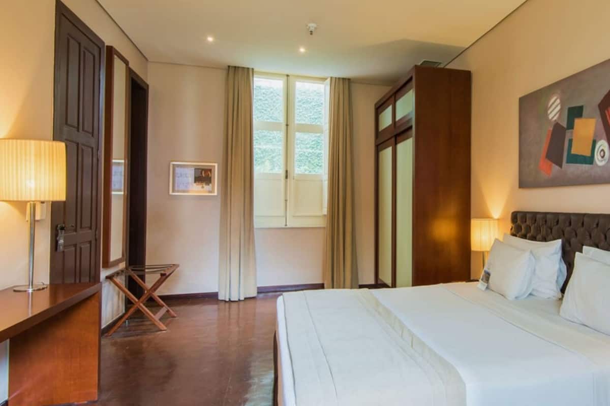 A hotel room with a large bed, a desk, a chair, a window, and a painting on the wall.