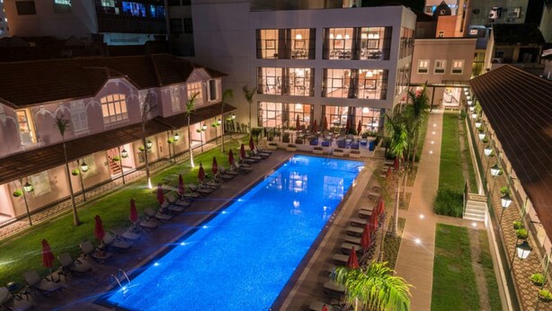 Una piscina con sillas de descanso y sombrillas, rodeada por un edificio bien iluminado con numerosas ventanas.