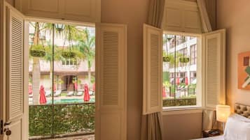 A hotel room with a bed, a nightstand, a lamp, and a view of an outdoor pool area with lounge chairs and umbrellas.