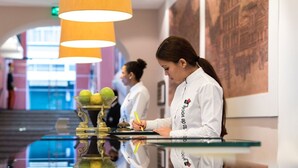 Zona de recepción con mostrador de cristal, un cuenco decorativo con frutas y una persona escribiendo en un bloc.