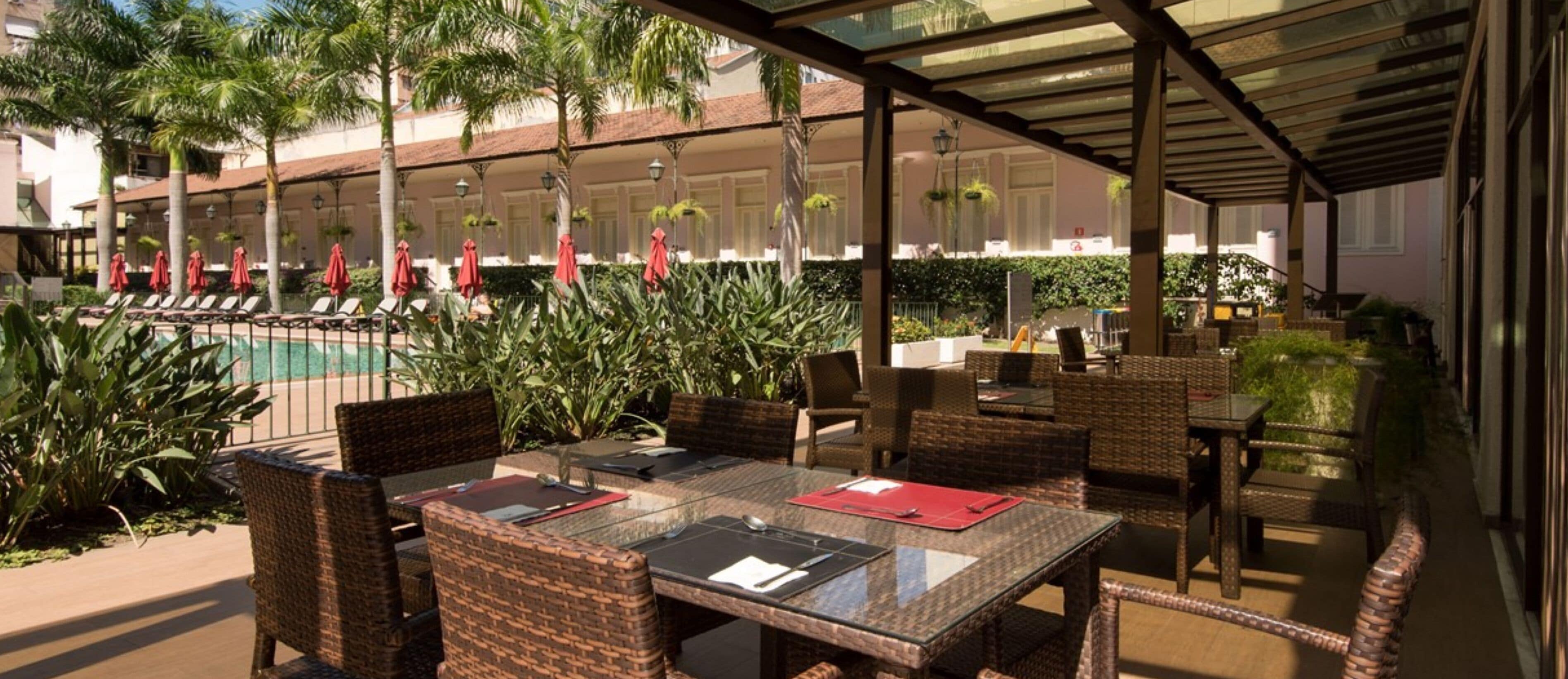 An outdoor dining area with wicker chairs and tables, a pool in the background, and palm trees.