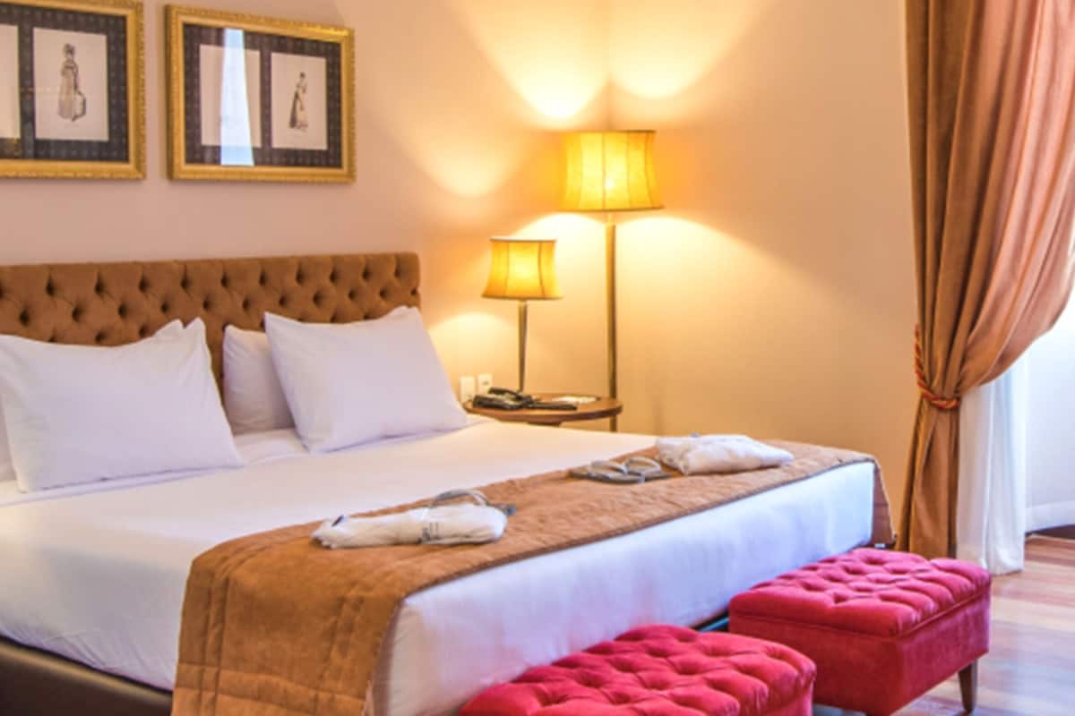 A hotel room with a large bed, two bedside lamps, a red ottoman, and framed artwork on the wall.