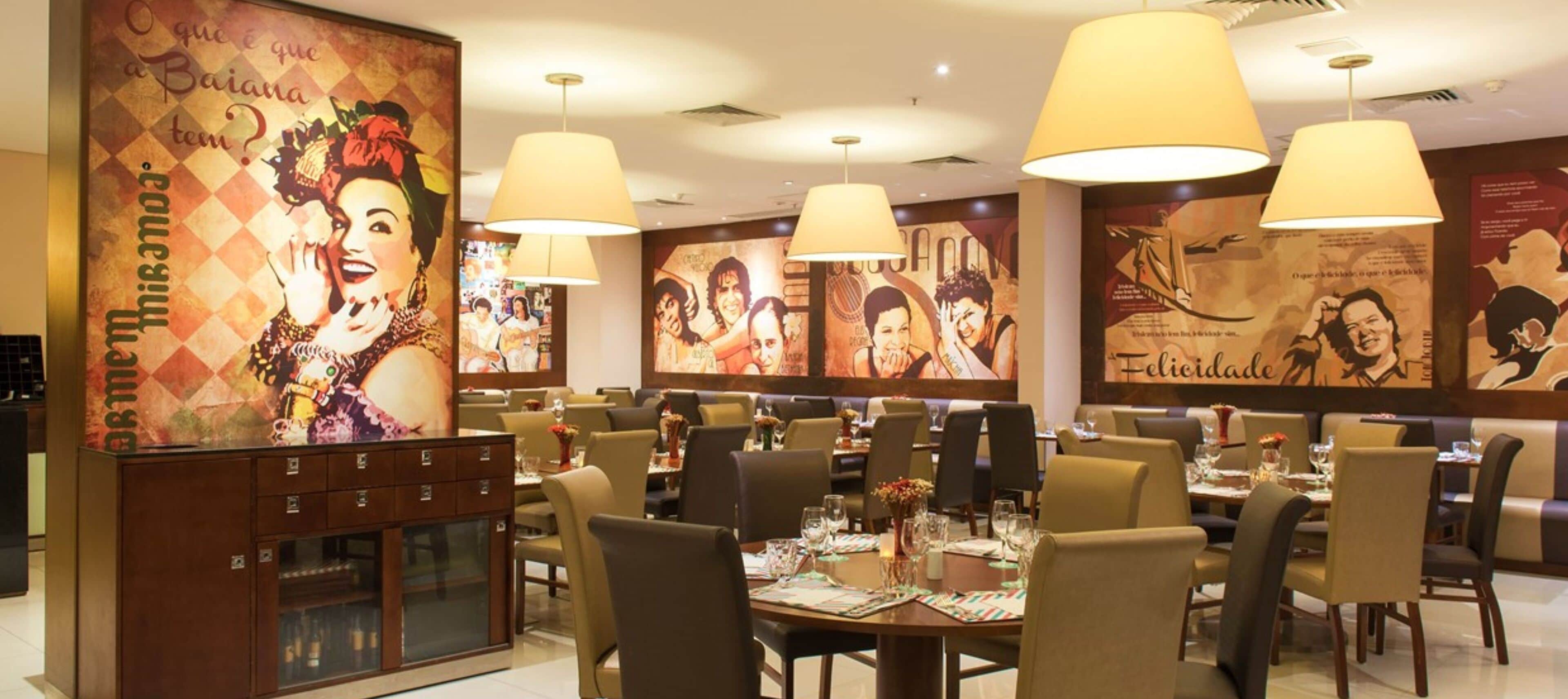 A restaurant with multiple dining tables and chairs, large hanging lamps, and a wall with various posters and artwork.