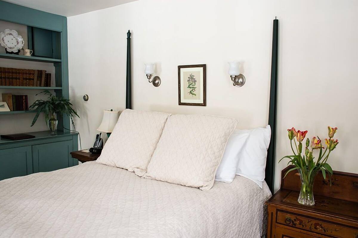 A neatly made bed with a quilted cover, a bedside table with a lamp, a wall-mounted picture, and a vase with flowers.