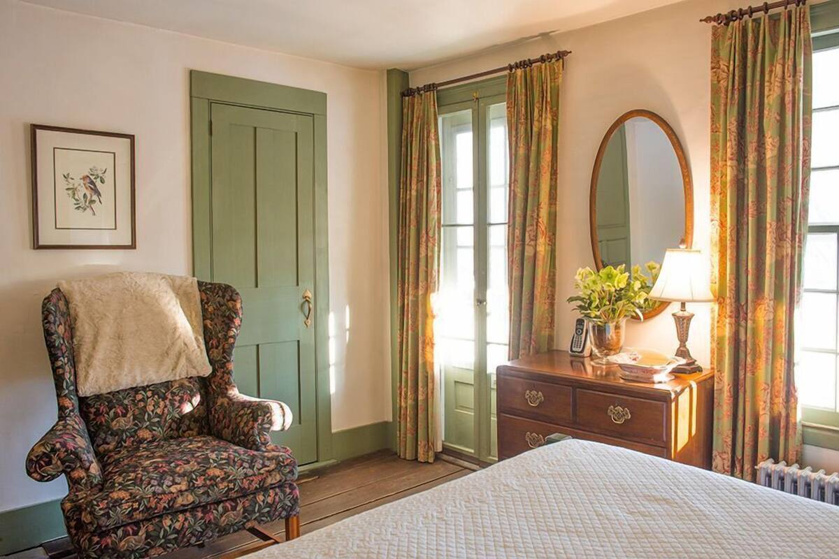 A bedroom with a floral-patterned armchair, a bed with a quilt, a dresser with a mirror, and green curtains.