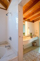 Un baño con ducha de azulejos blancos, techo de madera, lavabo con encimera de mármol y un espejo encima.