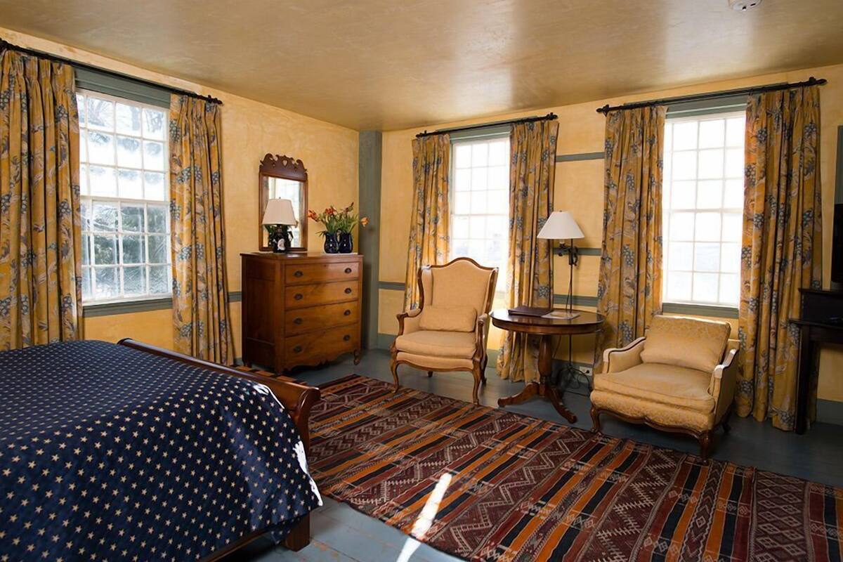 A hotel room with a bed, two armchairs, a wooden dresser, a small table, and a window with curtains.