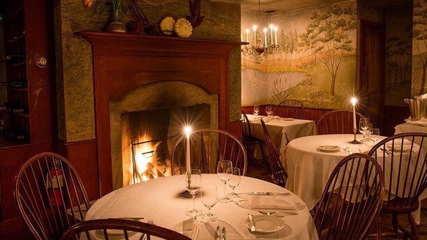 A dining area with round tables set for two, a fireplace, and a mural on the wall. - The Old Inn On The Green (Great Barrington)