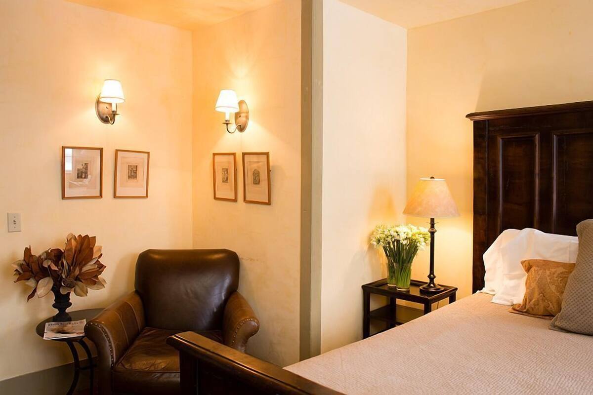 A hotel room with a bed, a chair, a nightstand with a lamp, and wall-mounted lights.