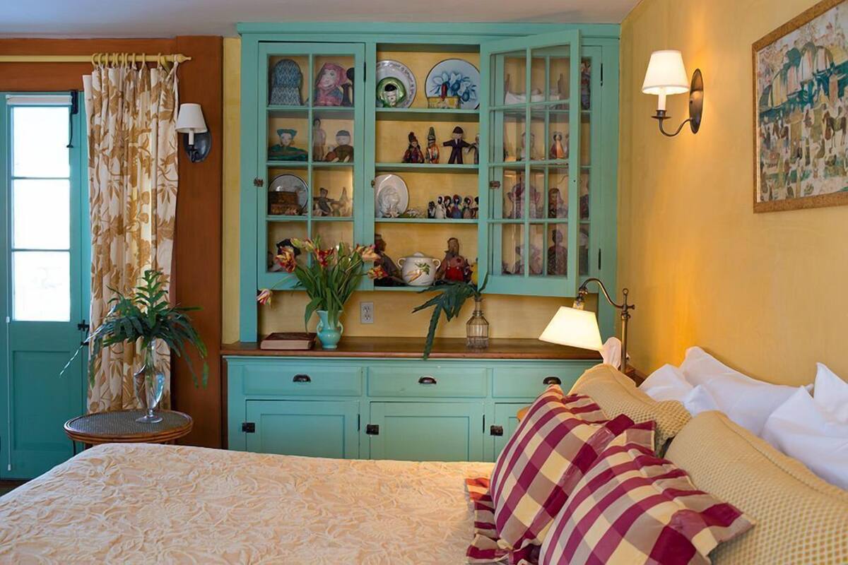 A bedroom with a bed, a teal cabinet with dishes and figurines, a lamp, and a painting on the wall.