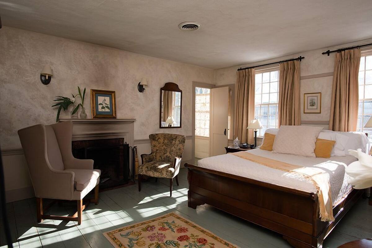 A bedroom with a large bed, a chair, a fireplace, and a window with curtains.