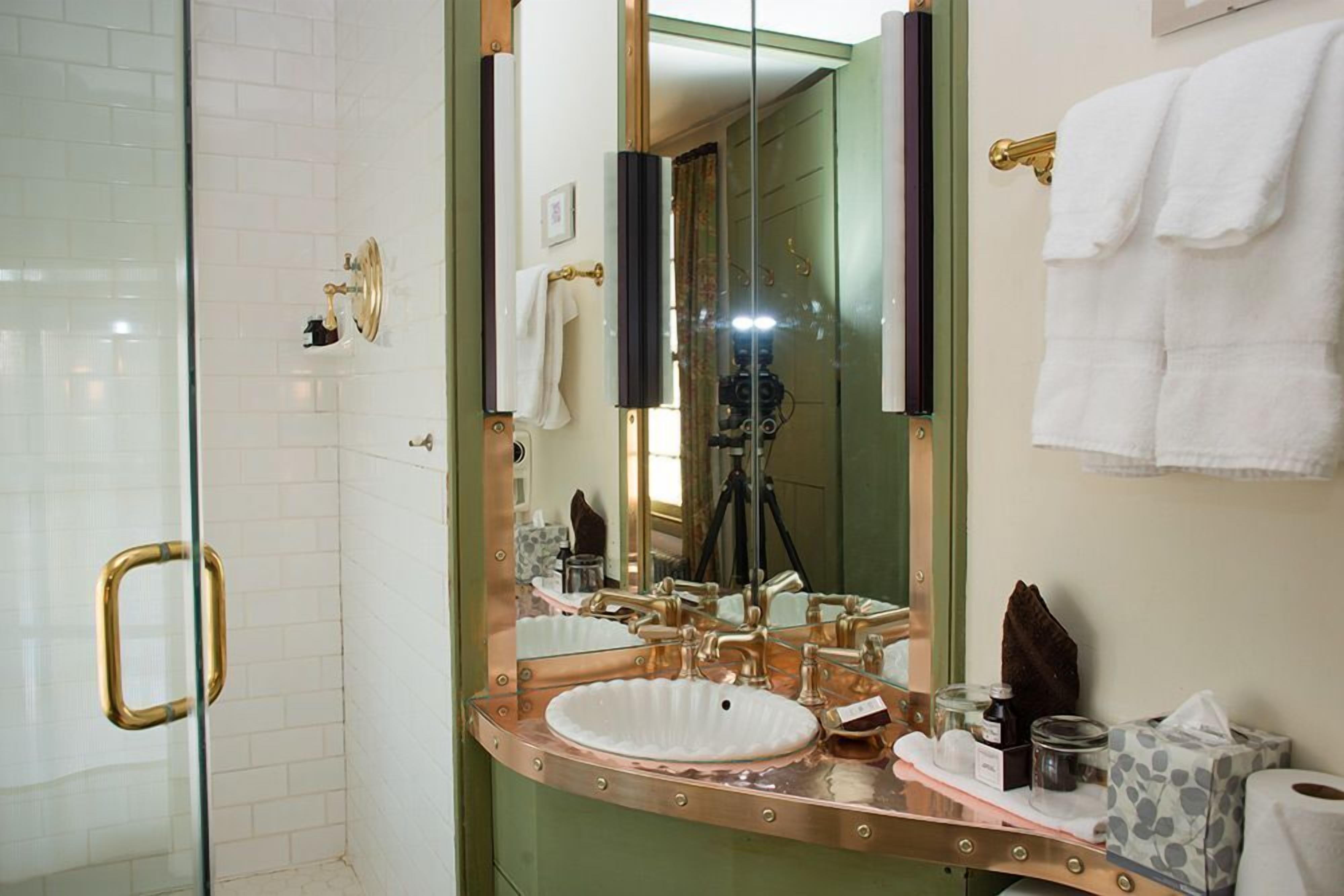 A bathroom with a glass shower door, a white sink with a copper faucet, and a large mirror.