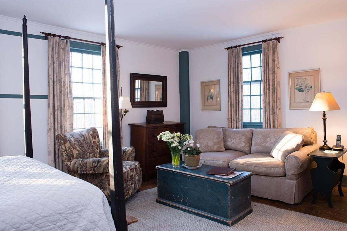 A bedroom with a four-poster bed, a sofa, a chair, a small table with a lamp, and a dresser.
