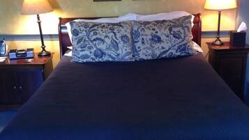 Cama com colcha azul escura e almofadas estampadas, com duas mesas de cabeceira laterais, uma delas com um telefone.
