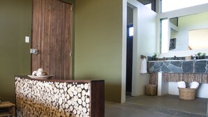 Ein Empfangsbereich mit einer hölzernen Bank, einer Wand aus gestapelten Holzscheiten, einer Holztür und einer Steinwand mit Waschbecken und Spiegel.