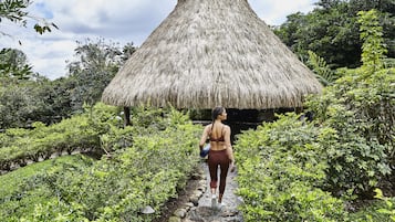 A person walking on a stone path towards a thatched-roof hut surrounded by lush greenery.